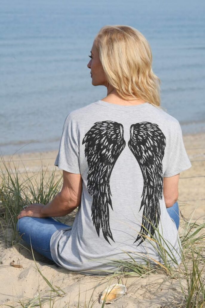 Dr. Madisen Harper, with angel wings t-shirt sits on a tranquil beach, reflecting near calm water.