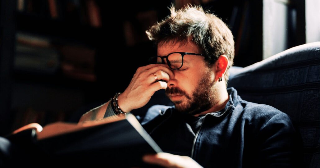 Man in contemplation, surrounded by books, expressing stress or fatigue in a cozy study.