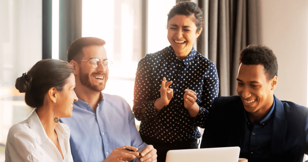 Collaborative diverse team sharing laughter around a laptop in a bright modern workplace.
